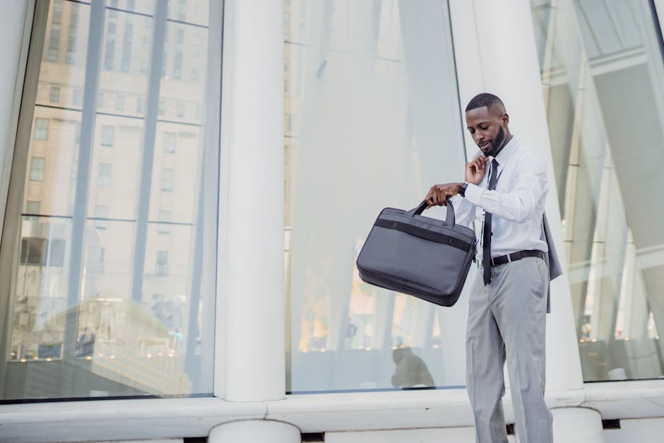 Professional man checking time with wristwatch while holding a laptop bag outside modern office.